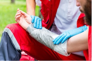 A first responder in a red uniform and blue latex gloves applying a large grey bandage to a person's forearm in an outdoor setting.