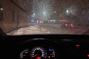 View from inside a car driving at night in heavy rain, with water droplets on the windshield and blurred city lights ahead.