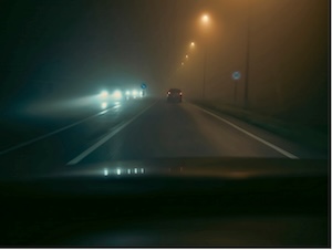 Nighttime highway driving in dense fog with low visibility, streetlights glowing and cars ahead with headlights on.