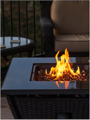 close-up shot of an outdoor gas fire pit table with a black granite top and a woven wicker base. Bright orange flames rise from a center burner filled with dark fire glass, reflecting on the polished table surface.