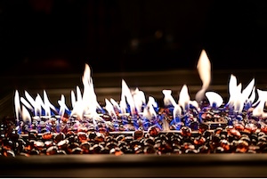 A long, rectangular outdoor fire feature at night. Small, flickering blue and white flames emerge from a bed of glowing red and blue glass fire beads.