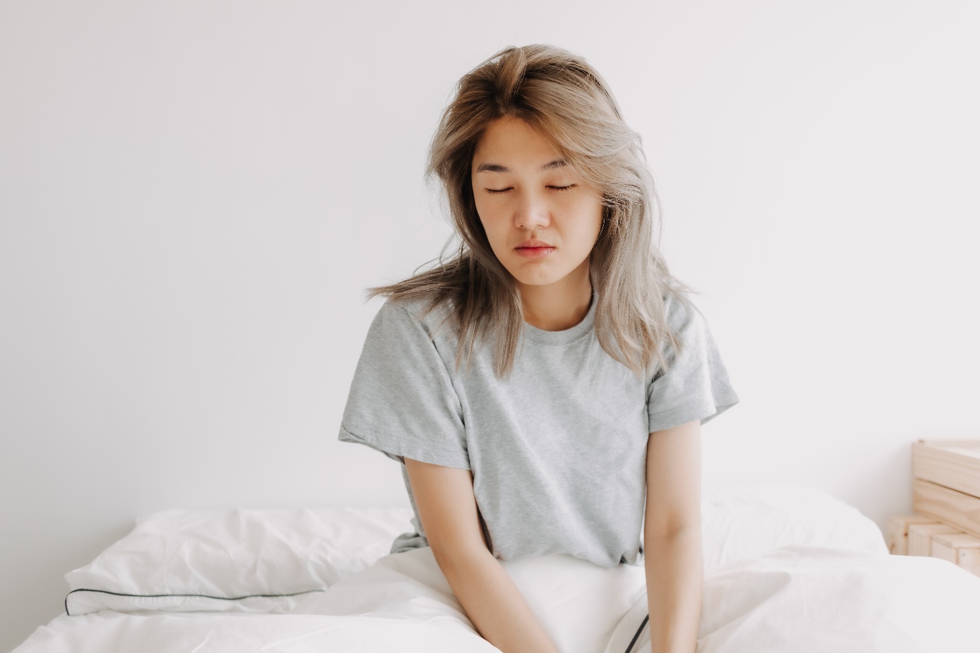 A young woman in a gray T-shirt sits on a white bed with eyes closed and shoulders slumped, conveying fatigue or sadness in a quiet bedroom.