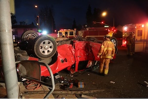 Overturned red car after a crash at night with firefighters and emergency vehicles responding at an intersection.